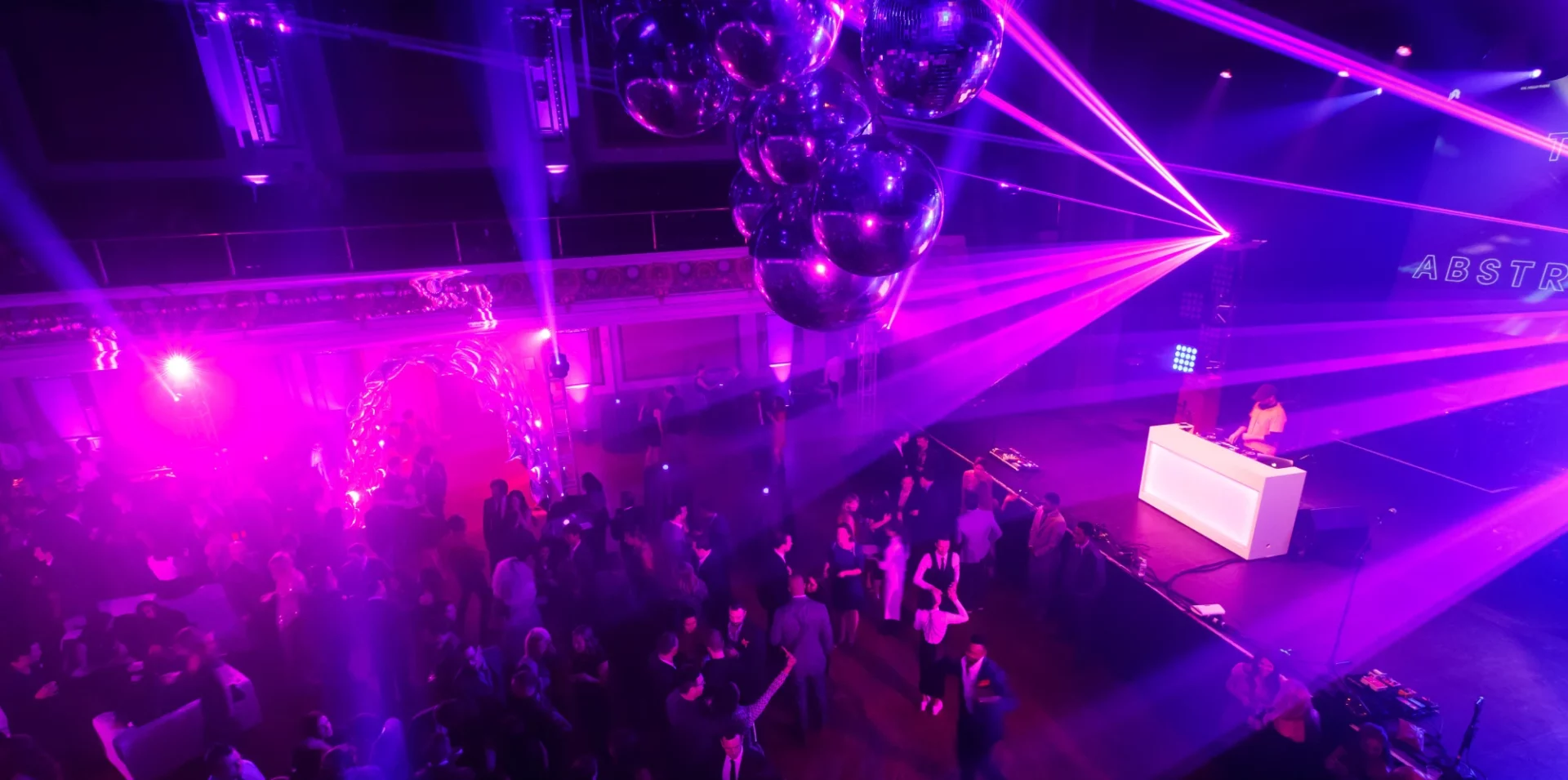 A lively nightclub party with a DJ, purple lighting, lasers, and people dancing.